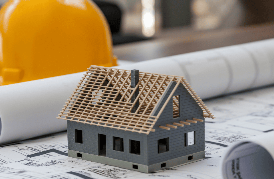 Image of a model house under construction, laying on a blueprint with a hardhat visible.